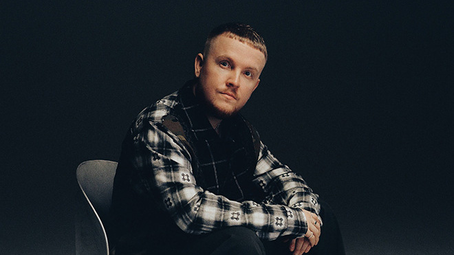 Chris Stassy assis sur une chaise dans un studio sombre